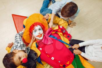 Funny clown and little children lying on the floor and pull their hands into the camera, top view. Birthday celebrating, baby holiday in playground. Childhood happiness, childish leisure