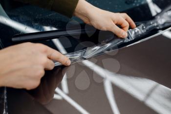 Male worker installs transparent protection film on car hood. Installation of coating that protects the paint of automobile from scratches. New vehicle in garage