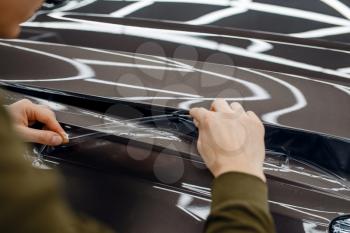 Male worker installs transparent protection film on car hood. Installation of coating that protects the paint of automobile from scratches. New vehicle in garage, tuning procedure