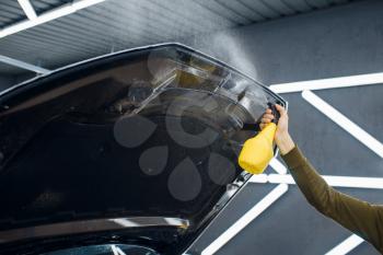 Male worker wets car hood surface with spray before applying of protection film. Installation of coating that protects the paint of automobile from scratches. New vehicle in garage, tuning procedure