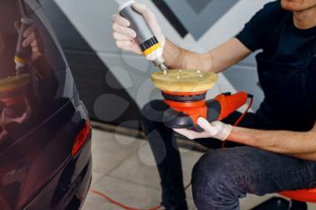 Male worker applies polish on polishing machine, car detailing. Preparation before installation of coating that protects the paint of automobile from scratches. Vehicle in garage, auto tuning