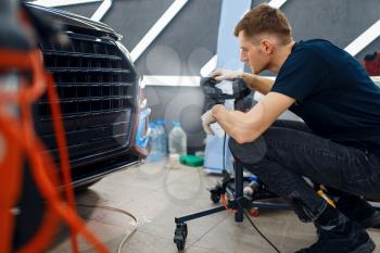 Male worker dries car protection film using a powerful lamp. Installation of coating that protects the paint of automobile from scratches. Vehicle in garage, auto tuning procedure