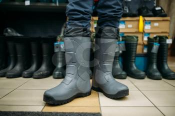 Fisherman tries on rubber boots in fishing shop. Equipment and tools for fish catching and hunting, accessory choice on showcase in store