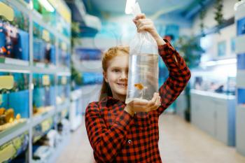 Happy little girl looking on goldfish in pet store. Child buying equipment in petshop