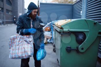 Male bearded beggar searching food in trashcan on city street. Poverty is a social problem, homelessness and loneliness, alcoholism and drunk addiction, urban lonely