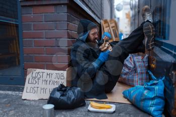 Bearded beggar eats burger on city street. Poverty is a social problem, homelessness and loneliness, alcoholism and drunk addiction, urban lonely