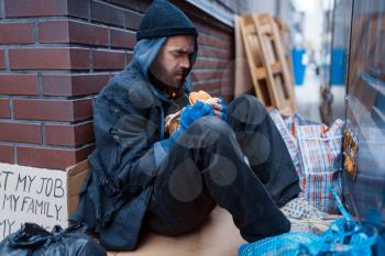 Bearded beggar eats burger on city street. Poverty is a social problem, homelessness and loneliness, alcoholism and drunk addiction