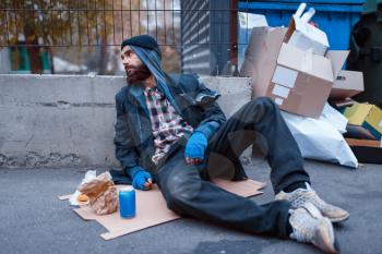 Bearded dirty beggar with food sitting at the trashcan on city street. Poverty is a social problem, homelessness and loneliness, alcoholism and drunk addiction
