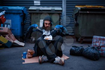 Bearded dirty beggar with knife and fork eats at the trashcan on city street. Poverty is a social problem, homelessness and loneliness, alcoholism and drunk addiction, urban lonely