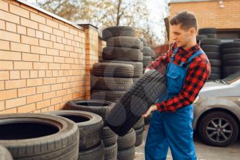 Male worker in uniform at the stack of tyres, tire service. Vehicle repair service or business, man repairing broken wheel