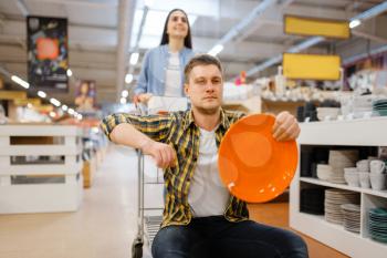 Young couple riding on cart in houseware store. Man and woman buying home goods in market, family in kitchenware supply shop