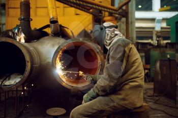 Welder in mask works with big metal pipe on factory, welding skill. Metalworking industry, industrial manufacturing of steel products
