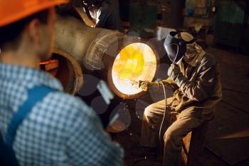 Welder in mask works with big metal pipe on factory, welding skill, worker in helmet on background. Metalworking industry, industrial manufacturing of steel products