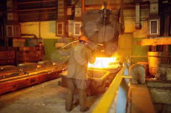 Steelmaker in helmet pours liquid metal from basket, steel factory, metallurgical or metalworking industry, industrial manufacturing of iron production on mill