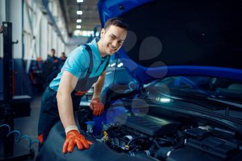 Smiling male worker in uniform checks vehicle engine, car service station. Automobile checking and inspection, professional diagnostics and repair