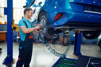 Worker in uniform removes wheel from vehicle on lift, car tire service station. Automobile checking and inspection, professional diagnostics and repair