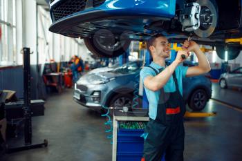 Worker in uniform repairing vehicle on lift, car service station. Automobile checking and inspection, professional diagnostics and repair