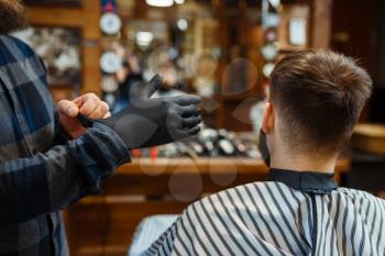 Barber puts on gloves, customer sitting in chair. Professional barbershop is a trendy occupation. Male hairdresser and client in hair salon