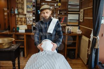 Barber and customer with towel on face, beard cutting. Professional barbershop is a trendy occupation. Male hairdresser and client in hair salon, pre-shave procedure