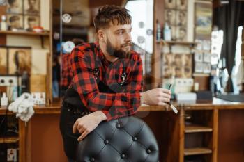 Barber with razor blade poses at the chair. Professional barbershop is a trendy occupation. Male hairdresser in retro style hair salon, accessories for cutting