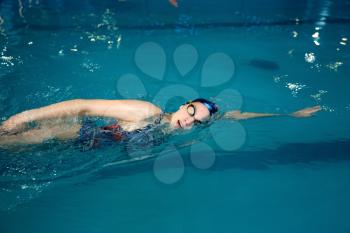 Female swimmer in swimsuit, cap and glasses swimming on her back in pool. The woman swims in the water, sport training