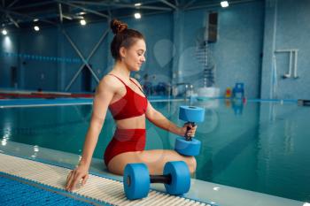 Female swimmer in red swimsuit poses at the poolside with dumbbells, water surface on background. The woman in pool, fitness workout, healthy lifestile, aqua aerobics