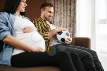 Husband with ball and his pregnant wife with belly joking at home, humor. Pregnancy, prenatal period. Expectant mom and dad are resting on sofa, health care