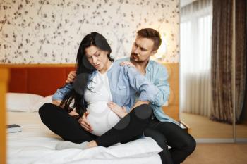 Husband doing massage to his pregnant wife with belly at home, bedroom interior on background. Pregnancy, prenatal period. Expectant mom and dad are resting on sofa, health care