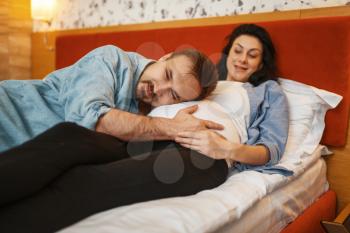 Husband listens to baby in pregnant wife's stomach at home, bedroom interior on background. Pregnancy, prenatal period. Expectant mom and dad are resting on sofa, health care