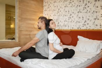 Happy couple, pregnant wife with belly and husband are sitting in yoga pose at home, bedroom interior on background. Pregnancy, prenatal period. Expectant mom and dad are resting in bed