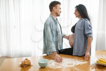 Happy couple, caring husband feeding his pregnant wife at home, kitchen interior on background. Pregnancy, prenatal period. Expectant mom and dad are resting in the morning