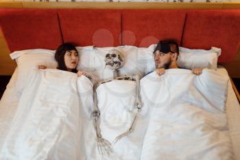 Husband, wife and human skeleton lying in the bed, bad relationship. Couple having a problems, family quarrel, conflict of married man and woman