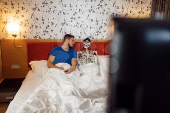 Man and human skeleton watching TV in the bed, bad relationship. Couple having a problems, family quarrel or conflict