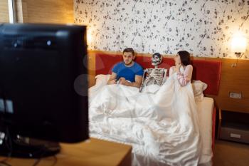 Husband watching TV, wife kissing human skeleton in the bed, bad relationship. Couple having a problems, family quarrel, conflict of married man and woman