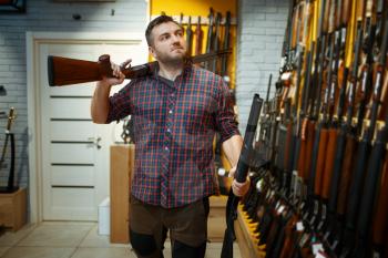 Man poses with two rifles at showcase in gun shop. Euqipment for hunters on stand in weapon store, hunting and sport shooting hobby
