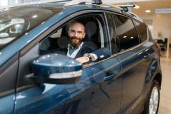 Smiling man poses in new automobile in car dealership. Customer in vehicle showroom, male person buying transport, auto dealer business