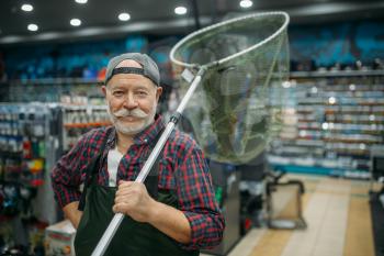 Male angler in rubber jumpsuit holds net in fishing shop, hooks and baubles on background. Fisherman buying equipment and tools for fish catching and hunting, assortment on showcase in store