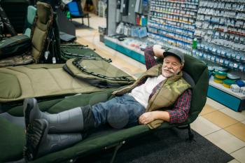 Smiling angler lying in sunbed in fishing shop, hooks and baubles on background. Equipment and tools for fish catching and hunting, accessory choice on showcase in store, bait assortment