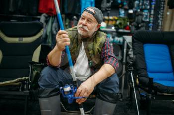Male angler in rubber boots holds rod in fishing shop, first-person view. Equipment and tools for fish catching and hunting, accessory choice on showcase in store