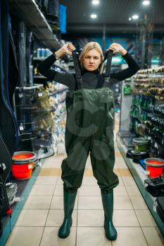Fisherwoman in rubber jumpsuit in fishing shop, hooks and baubles on background. Equipment and tools for fish catching and hunting, accessory choice on showcase in store