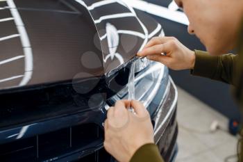 Male worker cuts transparent protection film on car hood. Installation of coating that protects the paint of automobile from scratches. New vehicle in garage, tuning procedure