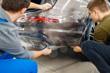 Two workers applies car protection film on rear bumper. Installation of coating that protects the paint of automobile from scratches. New vehicle in garage, tuning procedure