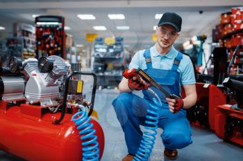 Male worker in uniform poses with pneumatic nailer in tool store. Choice of professional equipment in hardware shop, instrument supermarket