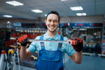 Smiling male worker in uniform holds measuring tape in tool store. Choice of professional equipment in hardware shop, instrument supermarket