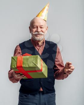 Happy elderly man in party cap holds gift box, grey background. Cheerful mature senior looking at camera in studio