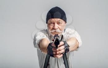 Elderly man poses with gun on grey background, gangster. Mature senior in hat holds weapon, robber in old age