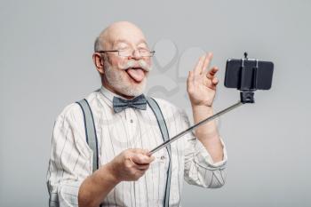 Happy elderly man makes selfie on phone, grey background. Mature senior showing his tongues at camera in studio