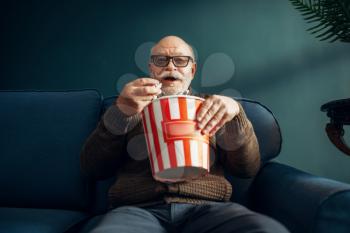 Elderly man with popcorn watching TV on couch in home office. Bearded mature senior poses in living room, old age businessman