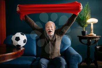 Elderly man with red scarf and ball watching TV, football fan. Bearded mature senior poses in living room, old age people leisures
