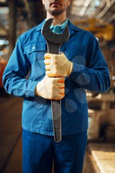 Male worker in uniform holds large wrench on factory. Industrial production, metalwork engineering, power machines manufacturing